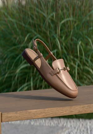 Mocassin slingback en cuir marron clair avec détail de boucle dorée, flottant au-dessus d'une surface en bois devant un fond d'herbe verte haute.