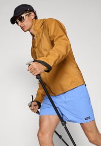 Lett brunt regnjakke med hette, kombinert med blå shorts, svart caps og solbriller, holder en svart turstang.