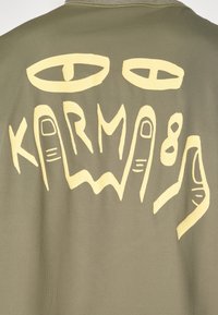Camiseta verde oliva con un gran estampado gráfico amarillo en la parte posterior, que representa ojos estilizados y la palabra "KARMA" con contornos de manos.