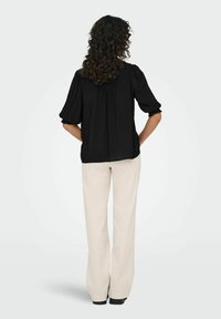 Sort kortærmet bluse med samlede detaljer, båret med cremefarvede vide bukser, vist fra ryggen mod en lys baggrund.