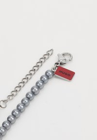 Grå pärlarmband med silverkedja, krokodilspänne och en röd rektangulär tagg med texten "HUGO" i svart.