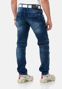 Dunkelblaue Jeans aus Denim mit schlanker Passform, ausgestattet mit abgenutzten Details, doppelter Naht und fünf Taschen. Tragen mit einem weißen Gürtel und hellen Sneakern.