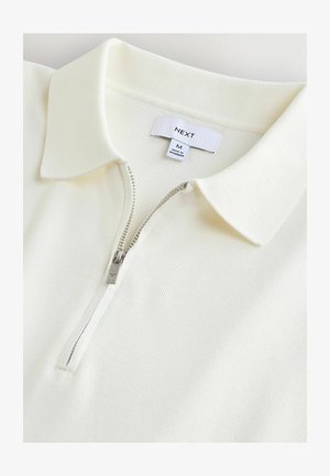White ribbed polo shirt with a zipper and collar. Features a silver zipper pull and a label reading "NEXT" and size "M". Smooth texture.