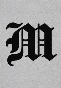 Lettera "M" nera ricamata su un tessuto grigio chiaro, caratterizzata da un design decorativo con curve intricate e accenti appuntiti.