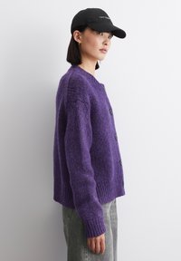 Cardigan lavorato a maglia viola con finitura testurizzata, dotato di scollatura rotonda, bottoni neri e polsini a costine. Indossato con jeans grigi e un cappellino nero.