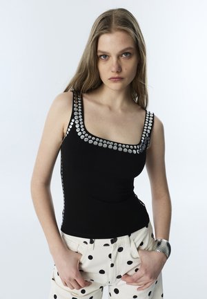 Giovane donna con lunghi capelli biondi indossa una canottiera nera con borchie argento e pantaloni bianchi con pois neri, mani nelle tasche.