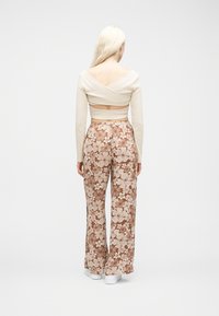 Γυναίκα με μακριά ξανθά μαλλιά που φοράει μπεζ τοπάκι με λουράκια στην πλάτη και χαλαρό καφέ και λευκό παντελόνι με floral σχέδιο, στέκεται μπροστά σε λευκό φόντο.