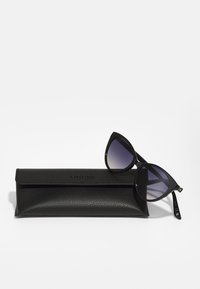Lunettes de soleil noires en forme d'œil de chat avec des verres dégradés, posées sur un étui noir texturé arborant un logo en relief. Design élégant et moderne.