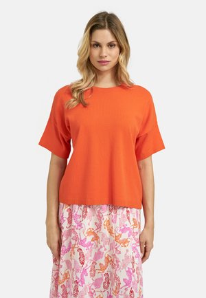 Woman with long blonde hair wearing an orange short-sleeve top and a skirt with pink and orange animal print patterns.
