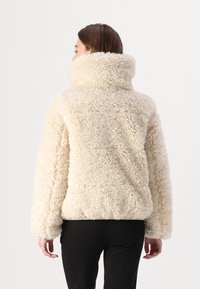 Kunstpelzmantel in Cremefarbe mit hohem Kragen und lockerem Schnitt. Verfügt über eine flauschige Textur und überschnittene Schultern, ergänzt durch schwarze Hosen.