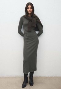 Lang, tætsiddende kjole i olivengrøn farve lavet af blødt stof, kombineret med et mørkebrunt tørklæde, der er draperet over skuldrene, og sorte ankelstøvler.