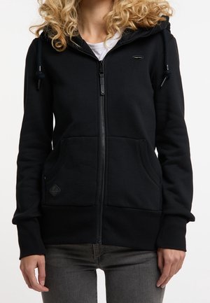 Femme portant un sweat à capuche noir zippé avec poches devant et cordons, associé à un jean gris foncé, vue des épaules aux hanches.