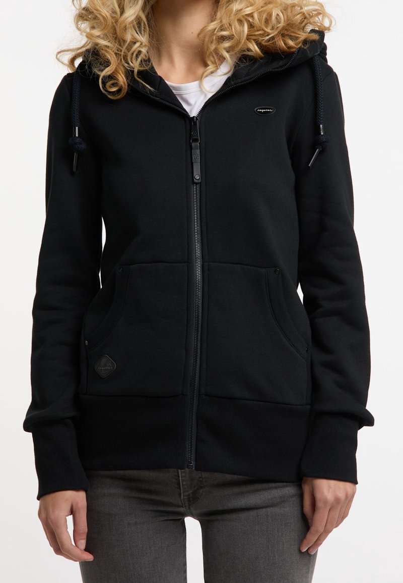 Femme portant un sweat à capuche noir zippé avec poches devant et cordons, associé à un jean gris foncé, vue des épaules aux hanches.