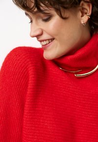 Rode gebreide trui met hoge col, voorzien van een textuurafwerking. Gouden hardware accent rond de hals complementeert de gedurfde kleur.