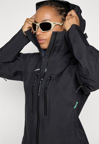 Black waterproof jacket with a hood, featuring multiple zippered pockets, textured fabric, and a green accent on the left side.