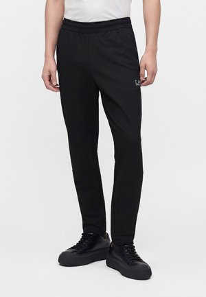 Mand iført sorte slim-fit joggingbukser og sorte snøresneakers, stående foran en ensfarvet baggrund.