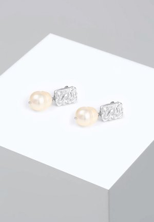 Pendientes de textura plateada que presentan grandes perlas redondas, adornados con pequeños acentos plateados. Exhibidos en una plataforma blanca minimalista.