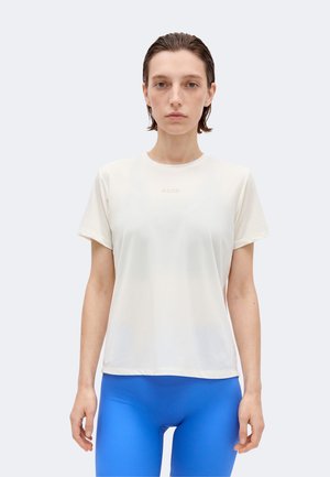 Γυναίκα με νωπά μαλλιά φοράει άσπρο κοντομάνικο μπλουζάκι με μικρό κείμενο και φωτεινά μπλε εφαρμοστά παντελόνια, στέκεται μπροστά σε ένα απλό φωτεινό φόντο.