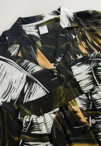 Camisa en blanco y negro con patrones de hojas, que incluye cuello con solapa, botones y una cintura fruncida. Tela suave y lisa con un brillo.