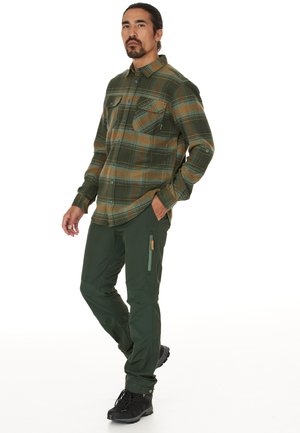 Groene en bruine geruite flanellen blouse met twee borstzakken, gecombineerd met donkere groene broek met een ritszak en versterkte knieën. Zwarte laarzen.