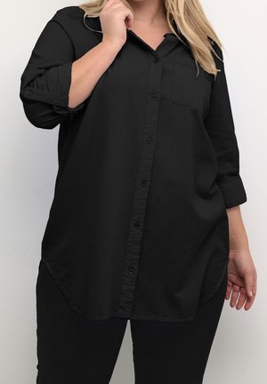 Femme portant une chemise noire ample à boutons avec manches retroussées et pantalon noir, main touchant le col, sur fond blanc uni.