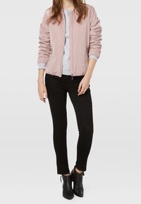 Lyserød bomberjakke med ribbede manchetter, parret med en grå sweater, sorte skinny jeans og sorte ankelstøvler foran en neutral baggrund.