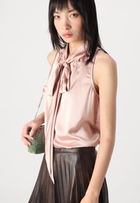 Blusa di satin rosa chiaro con dettaglio al collo legato, abbinata a una gonna plissettata marrone scuro e una piccola borsa a tracolla con gioielli verdi.