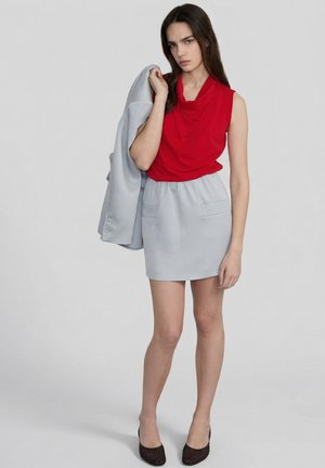 Kobieta w czerwonym topie bez rękawów i jasnoszarej spódnicy, trzymająca pasującą jasnoszarą marynarkę przewieszoną przez ramię, stojąca na jednolitym tle.