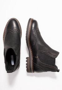 Pár černých kožených Chelsea bot s brogue detaily a hnědými prošitými podrážkami na bílém pozadí.