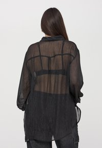 Blusa nera, trasparente, con maniche lunghe, dalla finitura testurizzata e un taglio rilassato. Presenta un colletto e un design con schiena aperta.