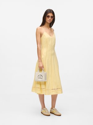 Woman wearing yellow midi dress and beige sneakers, holding a small white handbag, with sunglasses and straight hair.