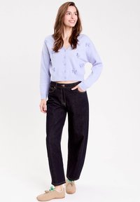 Ung kvinde smilende, iført en lyse lilla knappet cardigan med sløjfer, mørke vide bukser og beige sneakers med grønne snørebånd.