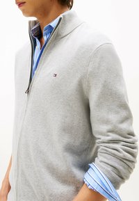 Suéter gris claro con cremallera y cuello acanalado. Presenta un pequeño detalle de logo y se combina con una camisa azul de rayas debajo del puño.