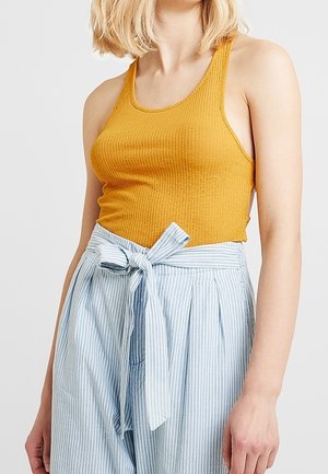 Vrouw draagt een mosterdkleurige geribbelde tanktop en hoog getailleerde lichtblauwe en witte gestreepte broek met een gestrikte tailleband.