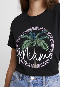 Sort bomulds T-shirt med et cirkulært design med et palmetræ og "Miami" i hvid skråskrift, omkranset af et græsk nøglemønster i lys pink.