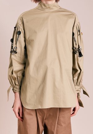 Blouse beige à col montant et manches longues avec poignets froncés et embellissements en perles florales noires sur les manches, portée avec un pantalon marron ample.