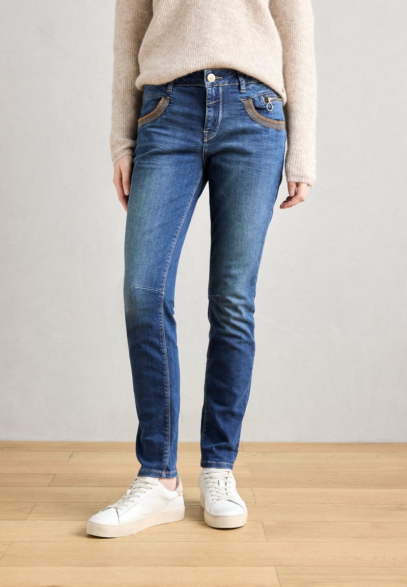 Jean bleu foncé coupe slim, avec poches avant soulignées de coutures contrastantes couleur beige et une poche zippée, associé à des baskets blanches.