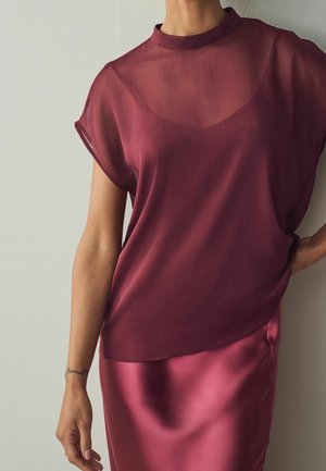 Vrouw draagt een doorschijnende bordeauxrode top over een mouwloze camisole en een bijpassende satijnen rok, staand tegen een effen lichte muur.