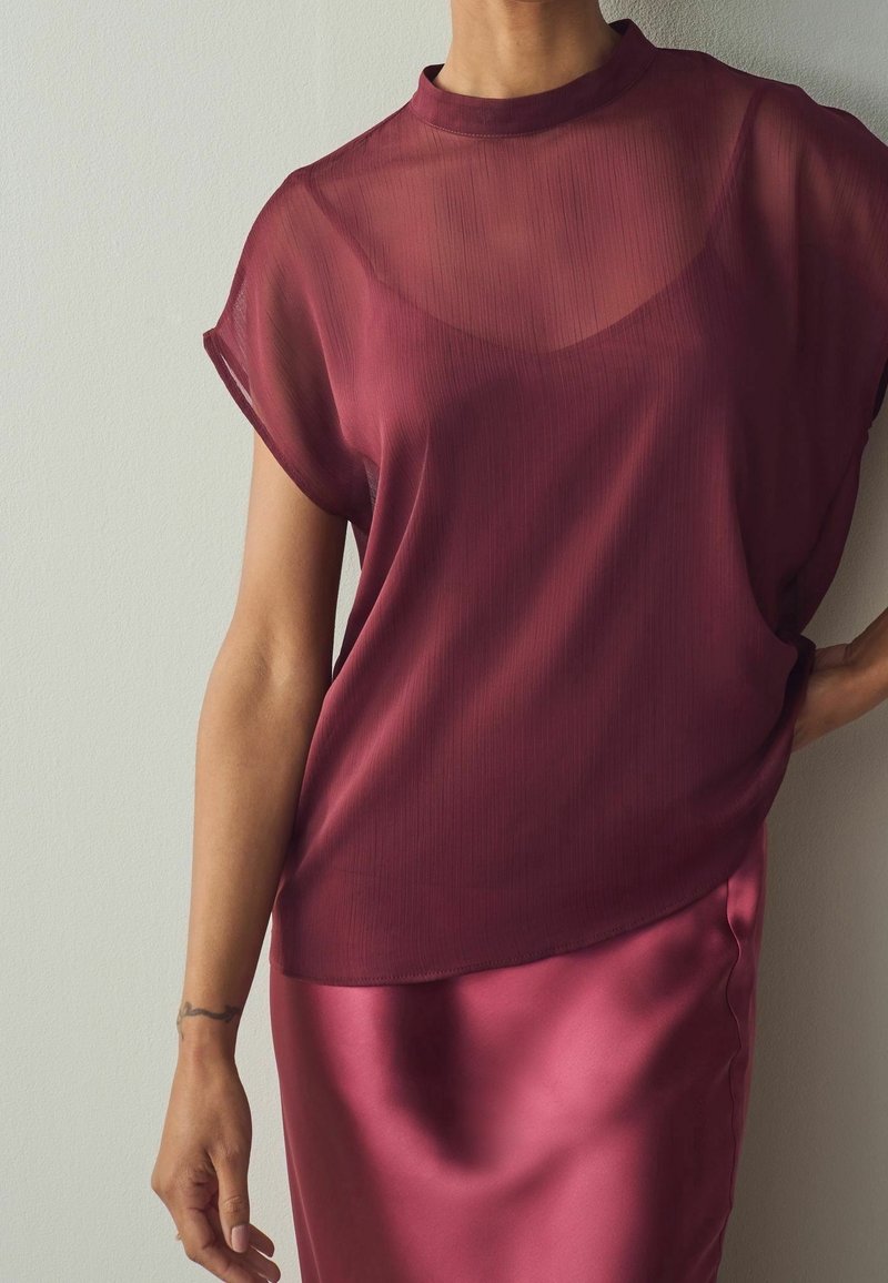 Femme portant un haut bordeaux transparent sur un caraco sans manches et une jupe en satin assortie, debout devant un mur clair uni.