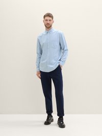 Light blue button-up shirt with a pocket, paired with dark navy trousers and black shoes. Model stands against a neutral background.