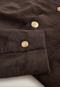 Brown corduroy fabric with vertical ridges, featuring gold-tone buttons with intricate designs on cuffs. Structured and textured surface.
