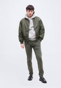 Νέος άνδρας που φοράει μπουφάν τύπου μπόμπερ ελιάς, παντελόνι που ταιριάζει, γκρι φούτερ με κουκούλα και μαύρα μοκασίνια, όρθιος με τα χέρια στις τσέπες του πάνω σε λευκό φόντο.