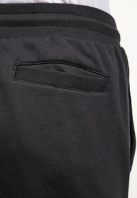 Detailní záběr na černé tepláky s elastickým pasem a horizontálními záhybovými kapsami na zadní straně na pravé straně.