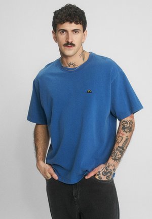 Mężczyzna z krótkimi ciemnymi włosami, wąsami i tatuażami, ubrany w niebieską koszulkę i czarne spodnie, stoi z rękami w kieszeniach.