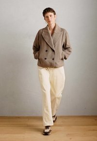 Personne marchant en avant portant une veste taupe à double boutonnage, un pantalon large crème et des baskets beiges sur un sol en bois.