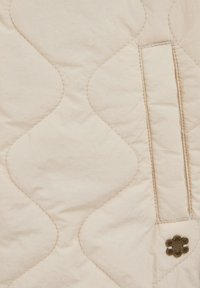 Prešívaná béžová látka s vlnitým vzorom, zdobená kovovým kvetom a skrytou kapsou na jednej strane, s jemnou textúrou.