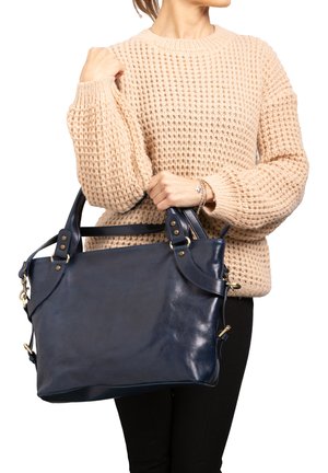 Persoon met een beige gebreide trui draagt een grote marineblauwe leren handtas met dubbele handvatten en messing beslag.