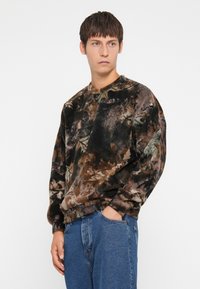 Bruine en zwarte sweatshirt met een patroon in zachte stof, voorzien van een ronde hals en lange mouwen. Gepaard met blauwe spijkerbroeken.