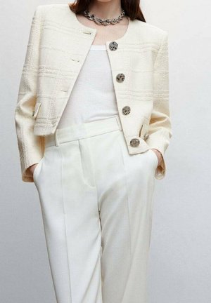 Femme portant une veste courte blanche texturée avec de gros boutons, un débardeur blanc côtelé, un pantalon blanc taille haute et un collier chaîne en argent.