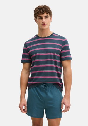 Mann trägt ein marineblaues T-Shirt mit horizontalen pinken Streifen und passende blaue Shorts mit Kordelzug, steht vor hellem Hintergrund.